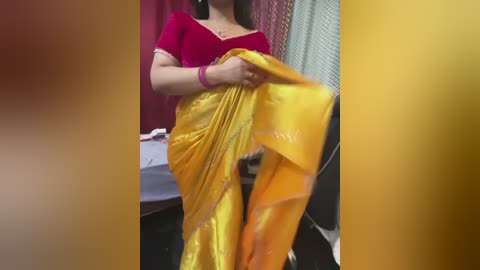 Video of a woman in a bright yellow sari, red blouse, and pink bangles, sitting on a chair, with maroon and white curtains in the background.