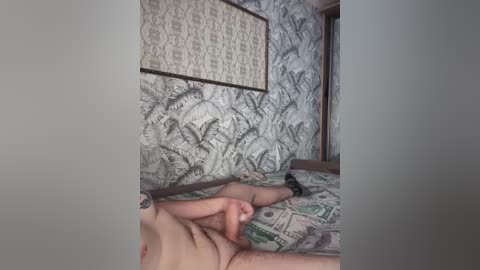 Video of a naked person with light skin, small breasts, and a tattoo on their right arm, lying on a bed with a green and white patterned duvet. The room features a wall with a large, intricate tapestry.
