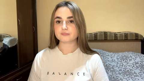 Video of a young woman with light skin, straight brown hair, and a nose piercing, wearing a white \"Balance\" t-shirt, standing in a beige bedroom with a bed and a mirror.