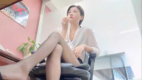 Video of an Asian woman with short black hair, wearing a sheer white blouse, black tights, and high heels, seated pensively on a black office chair. Background includes a pink wall, green plant, and abstract art.