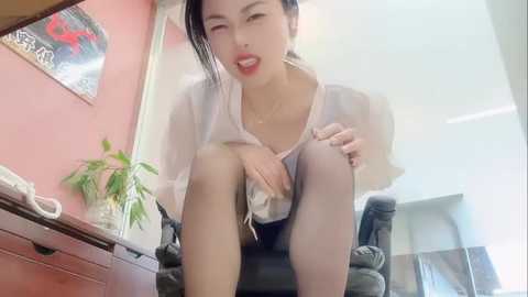 A video of an Asian woman with short black hair, sitting on a black chair in a brightly lit room, wearing a white blouse and black shorts, playfully licking her fingers.