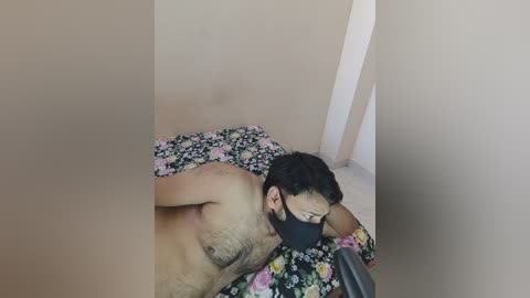 Video of a shirtless man with dark hair and beard, wearing a black mask, lying on a bed with a floral-patterned blanket. The room has beige walls and a vertical white pillar in the background.