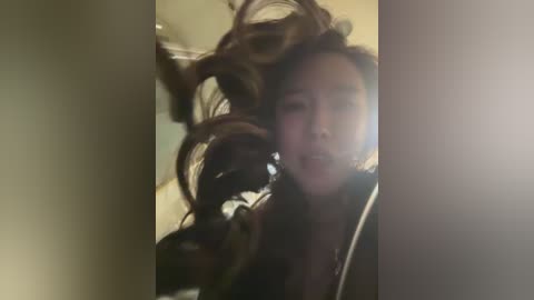 A blurry video captures a young woman with long, dark hair, her head tilted back, in a dimly lit room. Her hair is wild and windswept, creating a dynamic, chaotic effect.