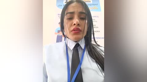 Video of a young woman with long black hair, wearing a white blazer, black tie, and blue lanyard. Background shows a blurred sign and other text.