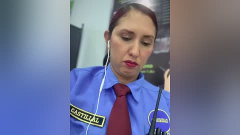 Video of a Latina female security guard in a blue uniform with a red tie, listening to headphones, standing in a blurred, indoor setting.
