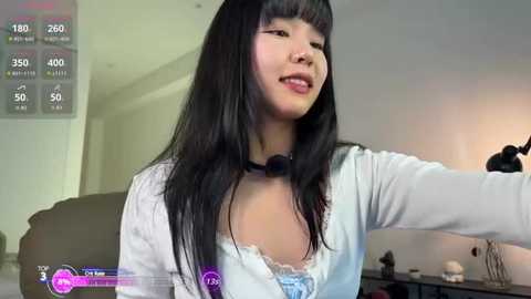 Video of an Asian woman with long black hair, wearing a white cardigan over a blue lace bra, smiling while playing a video game.