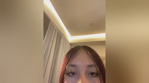 A video of a young woman with long, straight, red hair, slightly parted lips, and a neutral expression. She stands in a modern room with beige walls and a ceiling-mounted, rectangular, yellow light fixture.