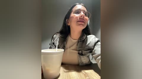 Video of a woman with glasses and long dark hair, wearing a denim jacket over a light shirt, seated at a wooden table with a white disposable cup.