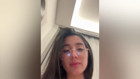 Video of a young woman with long dark hair, fair skin, and glasses, leaning against a beige wall with a modern, bright ceiling light.