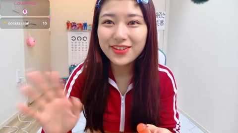 Video of a young Asian woman with long, straight dark hair, wearing a red zip-up jacket, smiling, holding a carrot, in a bright, clean room with a calendar on the wall.