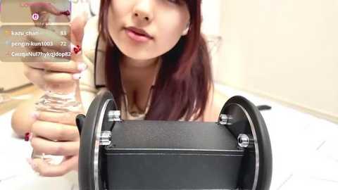 A video of a young woman with straight, dark red hair, holding a glass of water in one hand and a black microphone in the other, captured indoors with a blurred background.