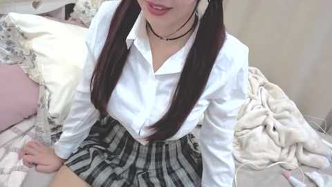 Video of an East Asian woman with long black pigtails, wearing a white blouse, black choker, and a black-and-white plaid skirt, sitting on a bed with white pillows and blankets.