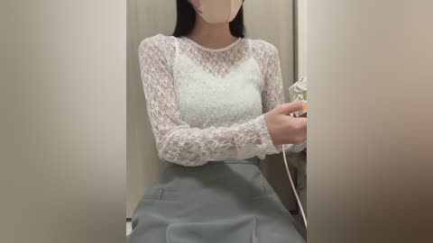 Video of an East Asian woman with long black hair, wearing a white lace top and grey skirt, standing in a beige-walled changing room, holding a white garment.