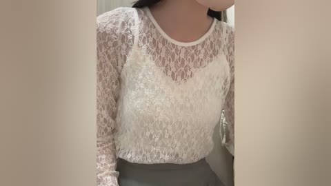 Video of a light-skinned woman with long dark hair, wearing a white lace top over a white bra, and grey pants. Background is blurred, featuring beige walls.