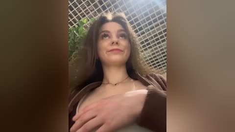 Video of a young woman with curly brown hair, wearing a brown cardigan, standing against a tiled wall. Her left hand rests on her chest, and she has a subtle smile.