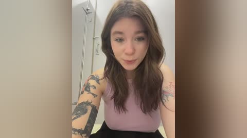 Video of a young Caucasian woman with long brown hair, wearing a sleeveless lavender top, showcasing detailed black ink tattoos on her arms. She's in a modern bathroom with white walls, a shower, and a brown door partially visible.