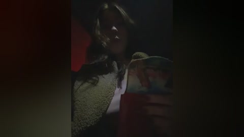 A dimly-lit video shows a woman with long, wavy brown hair, wearing a gray, textured coat, holding a colorful, patterned book. The background is dark and blurry, focusing on her face.