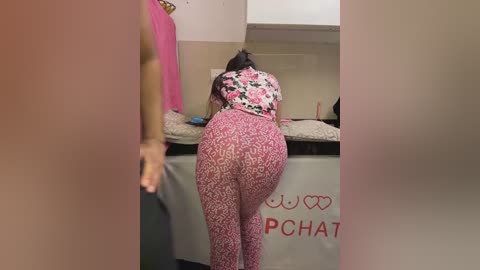 Video of a woman in a floral-patterned dress and leopard-print leggings, bending over to pick up something from a table in a cluttered, beige-walled room.