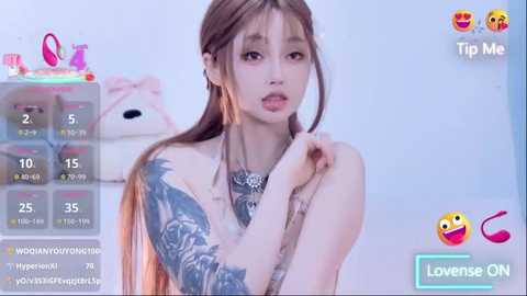 Video of a slender, light-skinned woman with long, straight brown hair and intricate tattoos on her arm. She has a youthful face with pink lips and large eyes, looking slightly confused. Background shows a soft, pastel room with a plush bear and a tip jar.