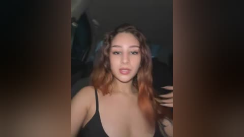 A video of a young woman with light skin, curly brown hair, and full lips, wearing a black spaghetti-strap top, taken indoors with dim lighting.