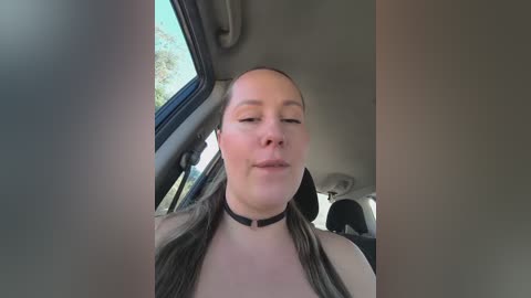 Media: A video of a fair-skinned woman with long brown hair, wearing a black choker, taking a selfie inside a car. The background shows the car's interior and a glimpse of greenery outside.