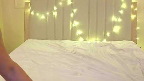 A video of a bed with white sheets and a beige headboard adorned with warm, yellow fairy lights, creating a cozy, intimate atmosphere. The background wall is plain and off-white.