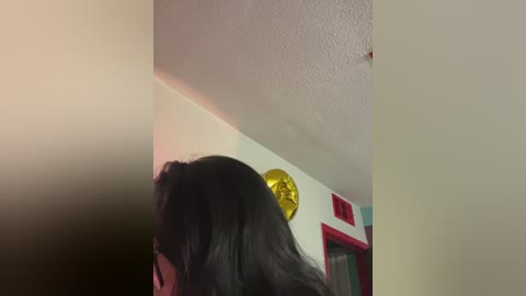 A video showing a woman with long black hair, viewed from behind, standing in a room with white walls and a golden sunburst wall decoration.