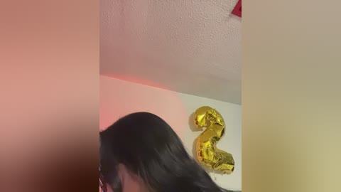 Video of a woman with long black hair, seen from the side, holding a gold foil number \"2\" balloon. Background includes a white wall and textured ceiling.