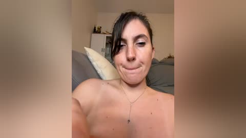 A candid video of a topless woman with light skin, brown hair, and a small nose ring, smiling in a bedroom with beige walls, gray pillows, and a white dresser.