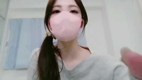 Video of a young Asian woman with long brown hair in a ponytail, wearing a light gray sweatshirt and a pink face mask, indoors with white walls and a blue curtain in the background.