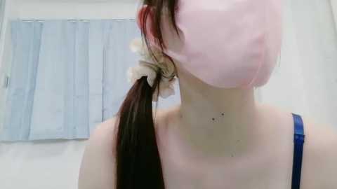 Video of a young woman with light skin, wearing a pink surgical mask and a blue spaghetti-strap top, her long brown hair tied in a high ponytail with a beige scrunchie. Background shows a light blue curtain.