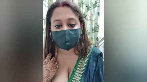 Video of a young woman with medium brown skin and straight brown hair, wearing a green face mask, traditional green and gold sari, and intricate henna on her hand.