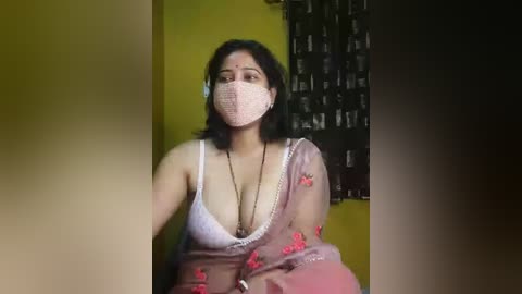 Video of a woman with medium skin tone, wearing a white lace bra, pink floral shawl, and face mask, seated against a green and black wall, with a blurred background.