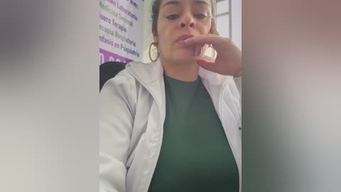 Video of a woman with medium skin tone, short hair, wearing a white lab coat over a green shirt, touching her chin pensively. Background shows blurred purple and white text.