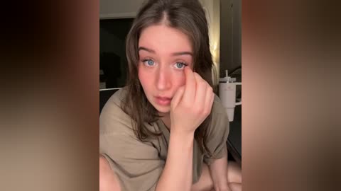 Video of a young woman with fair skin, blue eyes, and wavy brown hair, resting her chin on her hand in a thoughtful pose. She wears a beige t-shirt. Background features a dimly lit room with a door and furniture.