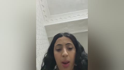 A video of a young woman with long, dark hair, light skin, and full lips, wearing a black top, standing in a tiled bathroom with white walls and a decorative ceiling.