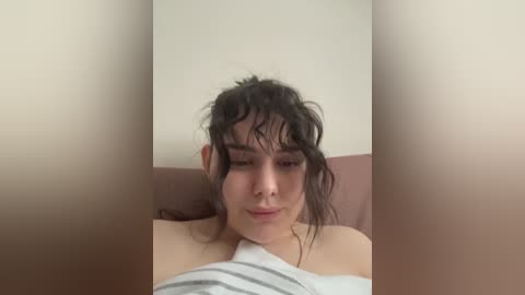 Video of a young woman with dark, messy hair, wearing a white shirt, lying on a bed against a plain, beige wall.