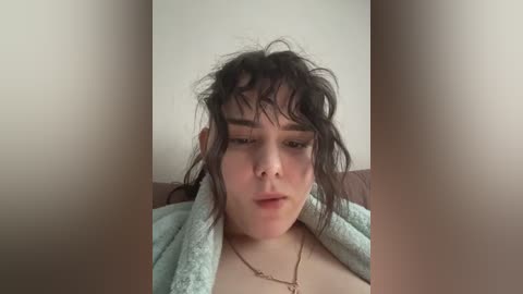 A video of a young woman with messy dark hair, wearing a green towel, looking tired, against a plain beige background.