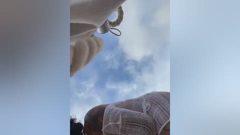 A low-angle video captures a person wearing a white knit sweater, partially visible from the chest down, against a bright, partly cloudy sky. The perspective emphasizes the sweater's texture and the sky's vastness.