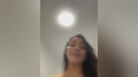 A blurry video of a young woman with long dark hair, wearing a bright orange top, standing in a dimly lit room with a bright ceiling light above.