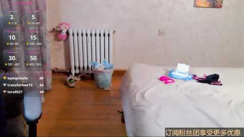Video of a messy bedroom with a bed, radiator, laundry basket, clothes, wooden floor, and a chair in the background.