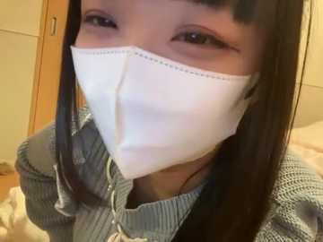 Video of a young Asian woman with long black hair and light skin, wearing a white surgical mask, a blue knit sweater, and a beige scarf, indoors with beige walls and a wooden door in the background.