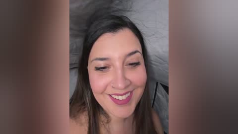 Media: Video of a smiling woman with long brown hair, fair skin, and pink lipstick, lying on gray bedding. Her expression is cheerful, and the image is framed with blurred edges.