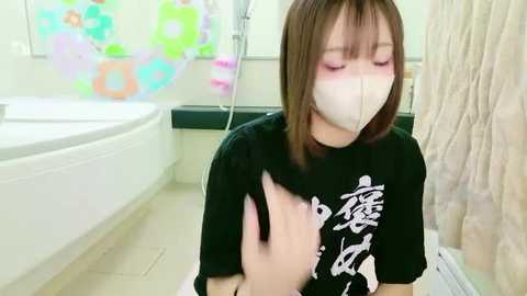 Video of a young woman with short brown hair and fair skin wearing a black T-shirt, face mask, and sitting in a modern, clean bathroom with a bathtub, colorful floral shower curtain, and beige towels.