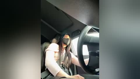 Video of a woman in a black face mask, wearing a beige top, driving a car. The interior is dimly lit with a dark grey dashboard visible.