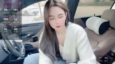 Video of a young Asian woman with long, straight brown hair, wearing a white cardigan, seated in a car, with a virtual reality display showing speed and location data.