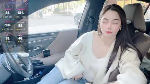 Video of a young Asian woman with long black hair, wearing a white sweater and blue jeans, resting her head on the car's armrest. The car's interior is modern, with a light-colored seat and a dashboard displaying a digital clock.