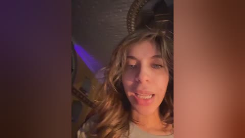 Video of a young woman with long, wavy brown hair, wearing a white shirt, smiling widely. Background features a dimly lit, textured metallic surface with a hint of blue light.