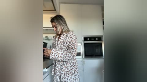 Video of a blonde woman in a leopard-print robe, cooking in a modern kitchen with white cabinets and stainless steel appliances.