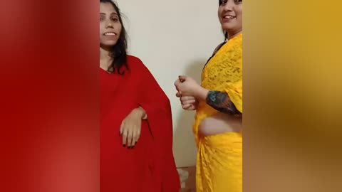 Video of two women in traditional Indian attire; one in a red long-sleeve dress, the other in a bright yellow outfit with floral designs. Both smiling, standing in a room with a plain white wall.